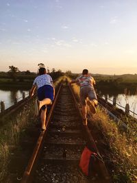 Rear view of male friends standing on railway bridge over river against sky during sunset
