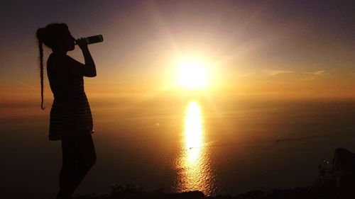 Silhouette woman drinking water at beach against sky during sunset