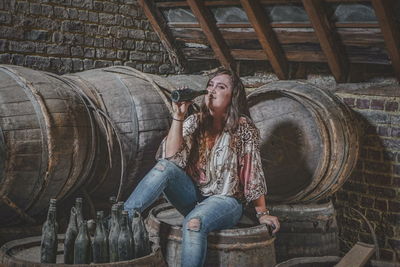 Portrait of smiling woman drinking alcohol from bottle while sitting on wooden barrel at abandoned cellar