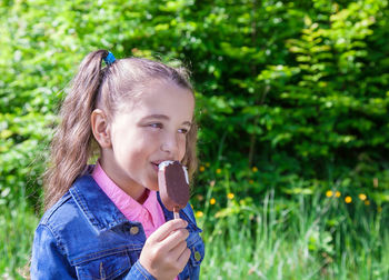 Side view of smiling girl eating popsicle standing by plants on field