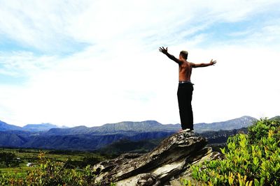 Low angle view of shirtless young man with arms outstretched standing on mountain against cloudy sky