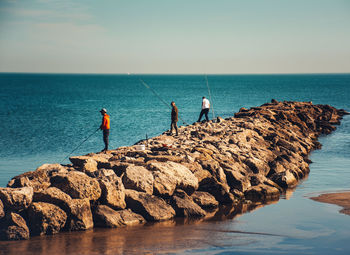 People fishing on rock by sea against sky