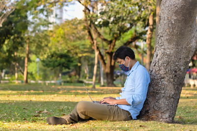 Side view of man wearing mask using laptop while sitting by tree trunk at park