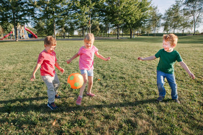 Preschool caucasian girl and boys friends playing soccer football on playground grass field outside. 