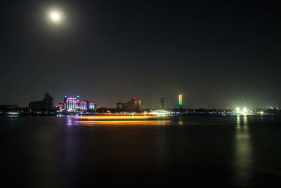 Illuminated buildings by river against sky at night