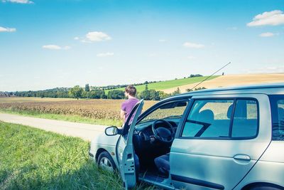 Man sitting on car hood against cloudy sky