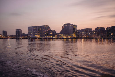 Illuminated buildings by sea against sky at sunset