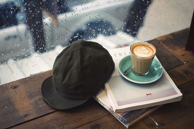 High angle view of coffee with books and cap on table by window in cafe