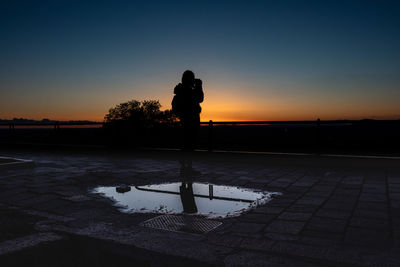 Silhouette of woman photographing while standing against sky during sunset