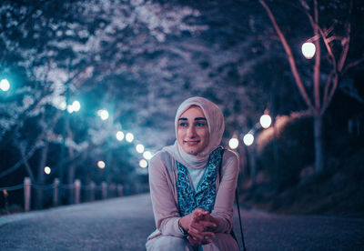 Portrait of smiling young woman standing against illuminated street at night