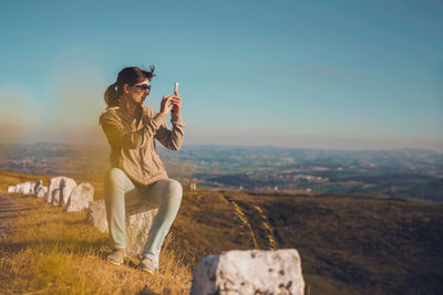 Mid adult woman wearing sunglasses photographing with smart phone while sitting on rock against blue sky