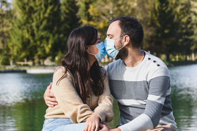 Young couple wearing masks sitting on bench by lake, kissing, new normal, covid, corona.