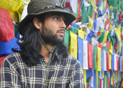 A bearded and long haired male tourist looking sideways while standing against tibetan prayer flags 