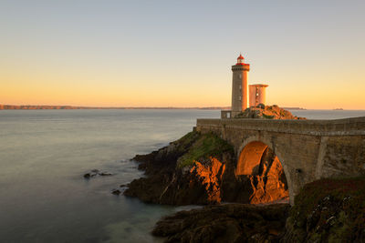 Lighthouse by sea against clear sky during sunset