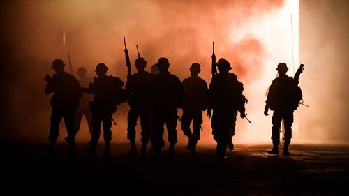 Silhouette people with weapons walking against smoke during sunset