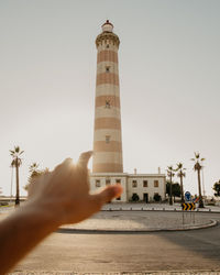 Hand gesturing towards lighthouse against clear sky