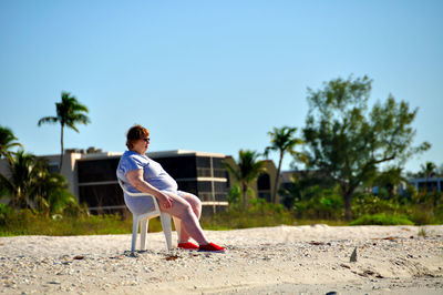 Side view of woman sitting against trees against clear blue sky