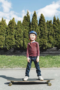 Full length portrait of happy boy standing on skateboard against trees