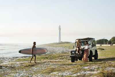 Man with surfboard walking towards female friends at beach against clear sky