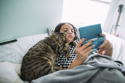 High angle view of girl playing video game while lying with cat on bed at home