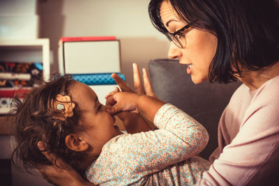 Mother talking to her small daughter who is pinching her nose and having fun at home.