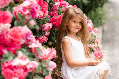 Cute baby girl 5-6 year old hold rose flower sit over bloom bushes in garden outdoor. spring season