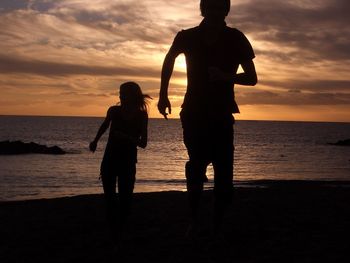 Silhouette brother and sister walking at beach against sky during sunset