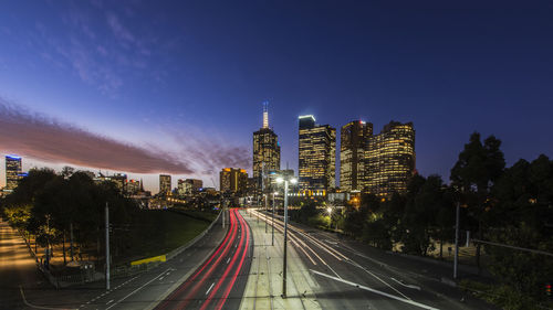 Light trails on road amidst buildings against sky at night