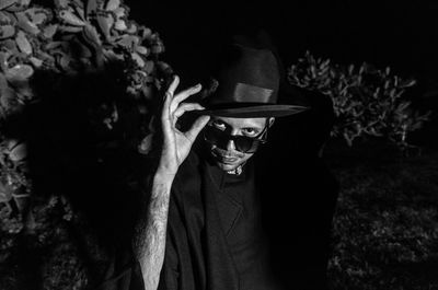 Portrait of young man wearing hat and sunglasses against plants at night