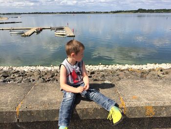 Boy looking away while sitting on retaining wall by sea