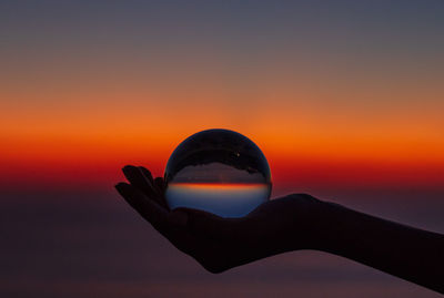 Cropped hand holding crystal ball against sky during sunset