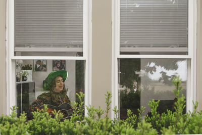 A woman sits singing at a window playing guitar in living room at home