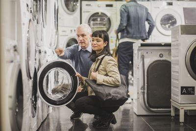 Mature owner pointing to washing machine while female customer crouching in electronics store