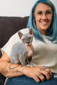 Cheerful woman with dyed hair and sphynx cat looking at camera while resting on couch at home