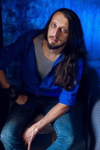 Man with long hair and beard sits at home on a chair by the window in a blue shirt and jeans