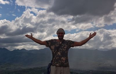 Portrait of smiling man with arms outstretched standing by mountains against sky