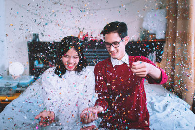 Cheerful couple throwing confetti while sitting on bed at home