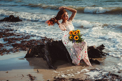 Full length of woman with flowers sitting on rock at sea shore against sky