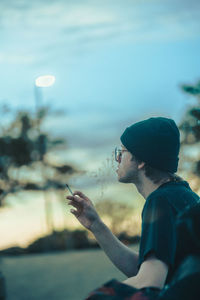 Side view of man holding a smoke against sky during sunset