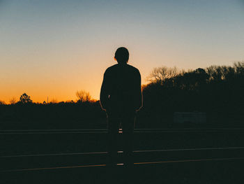 Silhouette man standing amidst railroad tracks on field against sky during sunset