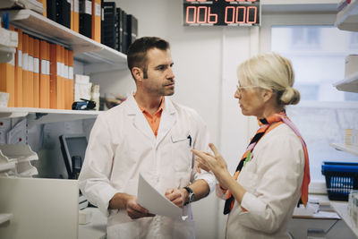 Mature medical colleagues discussing over documents by rack at pharmacy store