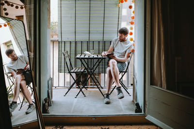 Young redhead male enjoys reading in his balcony sitting outdoors. he holds a book while reading 