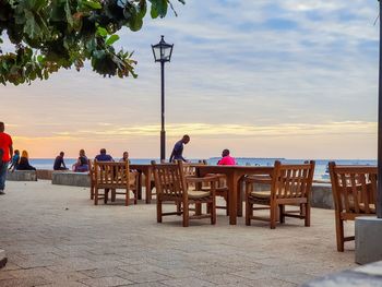 People sitting on table at beach against sky during sunset