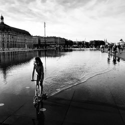 Woman on bicycle by river in city against sky