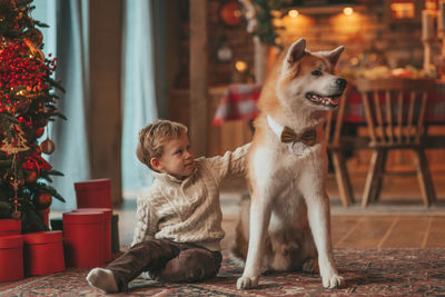 Candid authentic happy little boy in knitted beige sweater hugs dog with bow tie at home on xmas