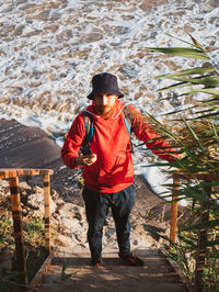 Bearded man in red hoody with backpack standing against sea with reeds. millennial guy travel alone.