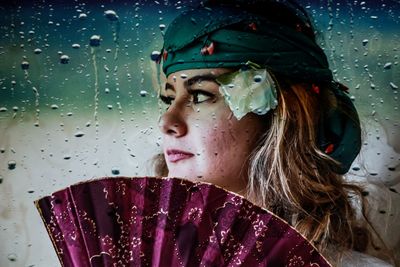 Close-up of young woman with folding fan seen through wet glass window