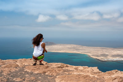 Rear view of mid adult woman looking at sea while crouching on mountain against sky