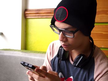 Young man with headphones wearing knit hat using mobile phone while sitting against wall