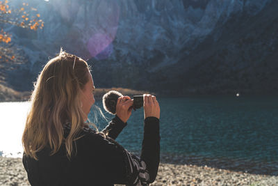Woman standing in front of beautiful mountain lake filming with video camera as sun begins to set